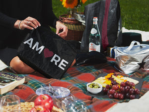 Woman holding black with "AMORE" zipper pouch surrounded by picnic foods