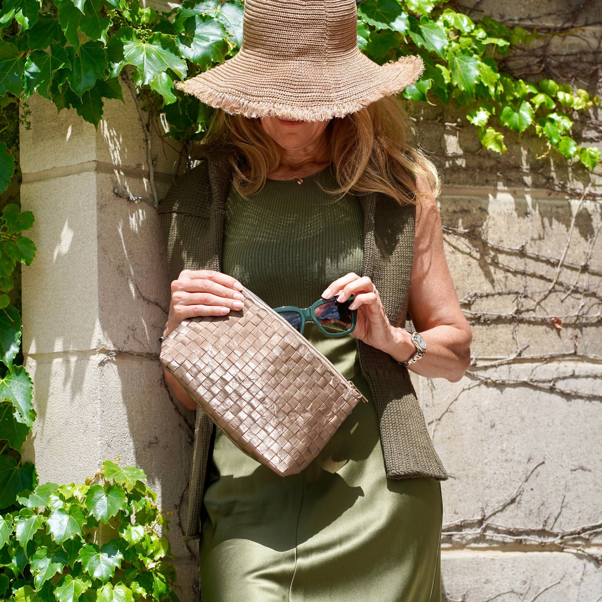 Woman wearing hat and army green dress pulling sunglasses out of woven shimmer bronze clutch