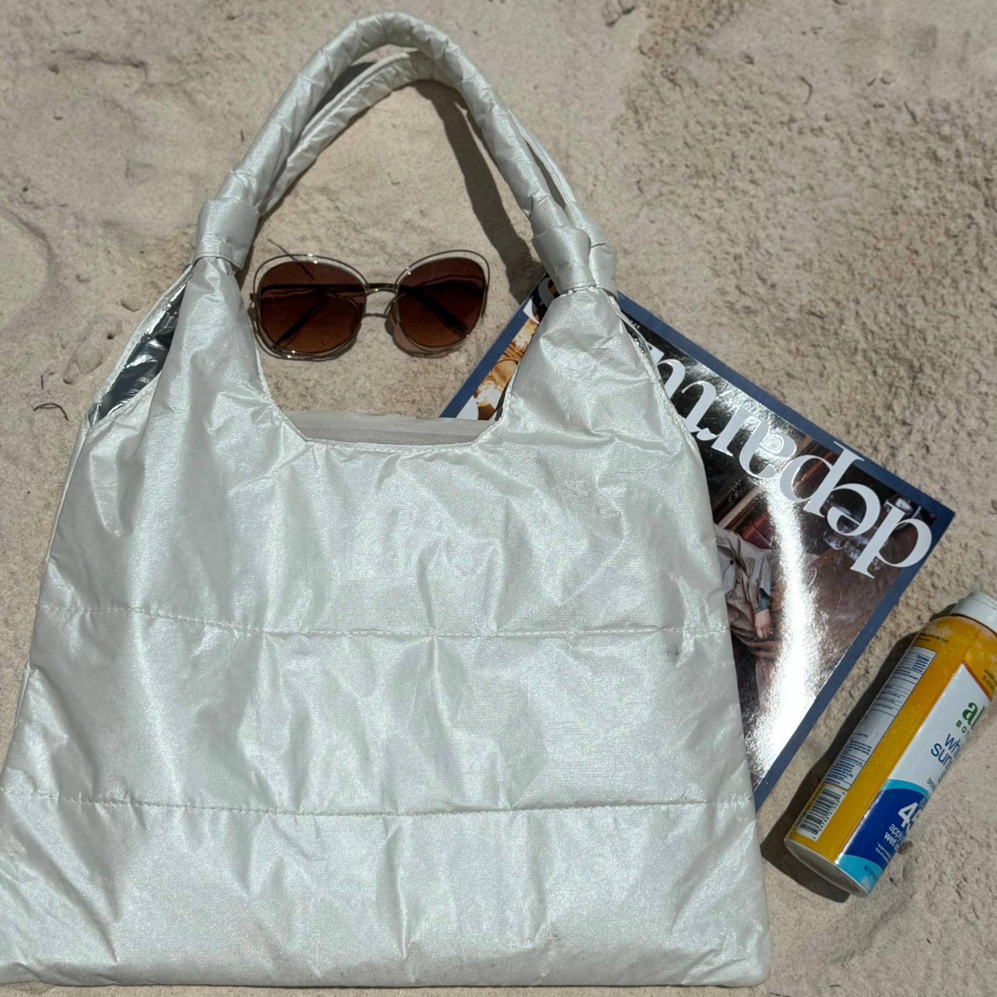 Shimmer white puffer tote bag laying in sand with sunglasses, magazine, and sunscreen