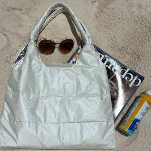 Shimmer white puffer tote bag laying in sand with sunglasses, magazine, and sunscreen