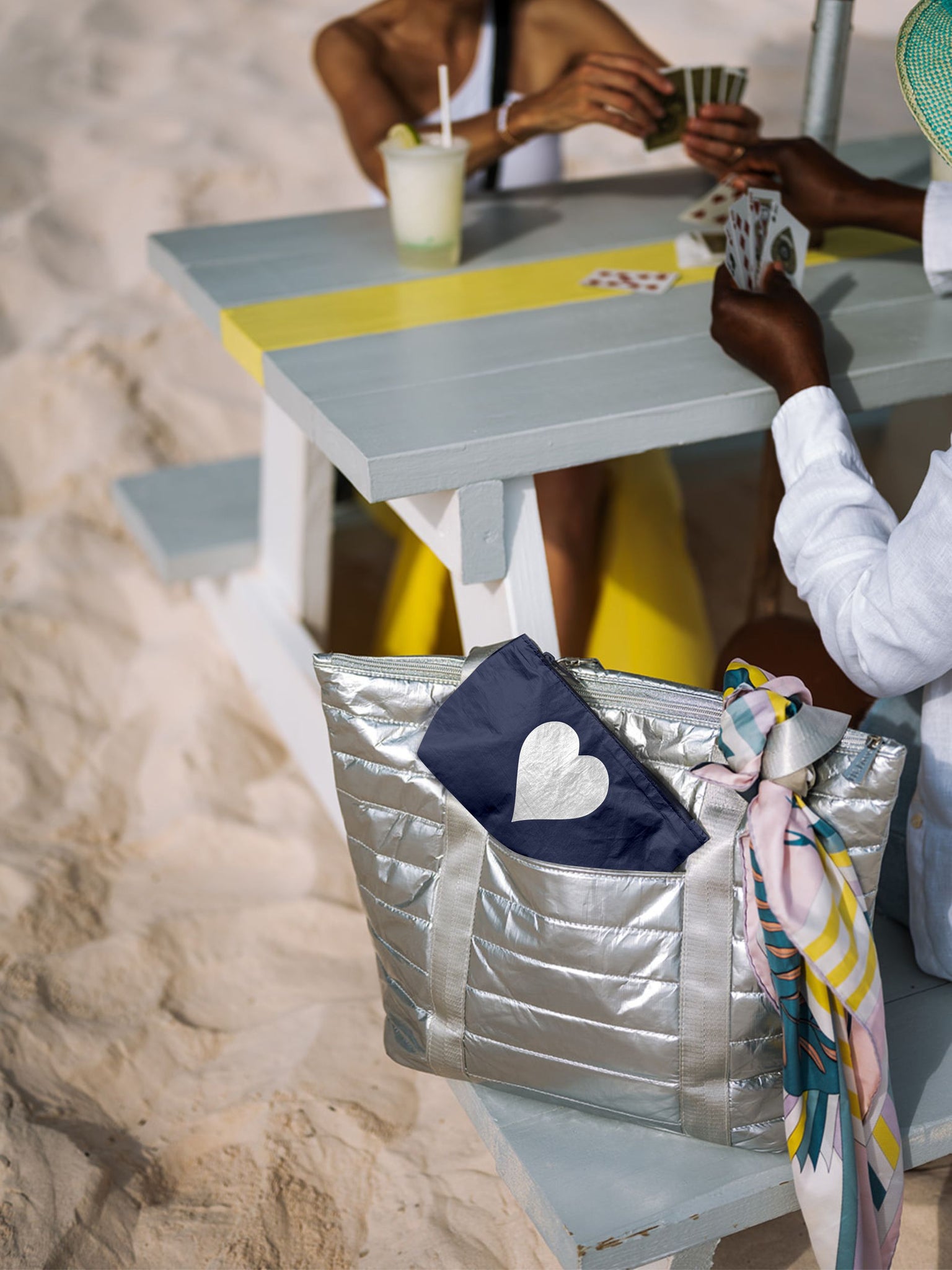 Silver puffer tote with a shimmer navy mini pouch sitting on a bench on the sand