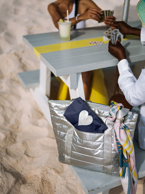 Silver puffer tote with a shimmer navy mini pouch sitting on a bench on the sand