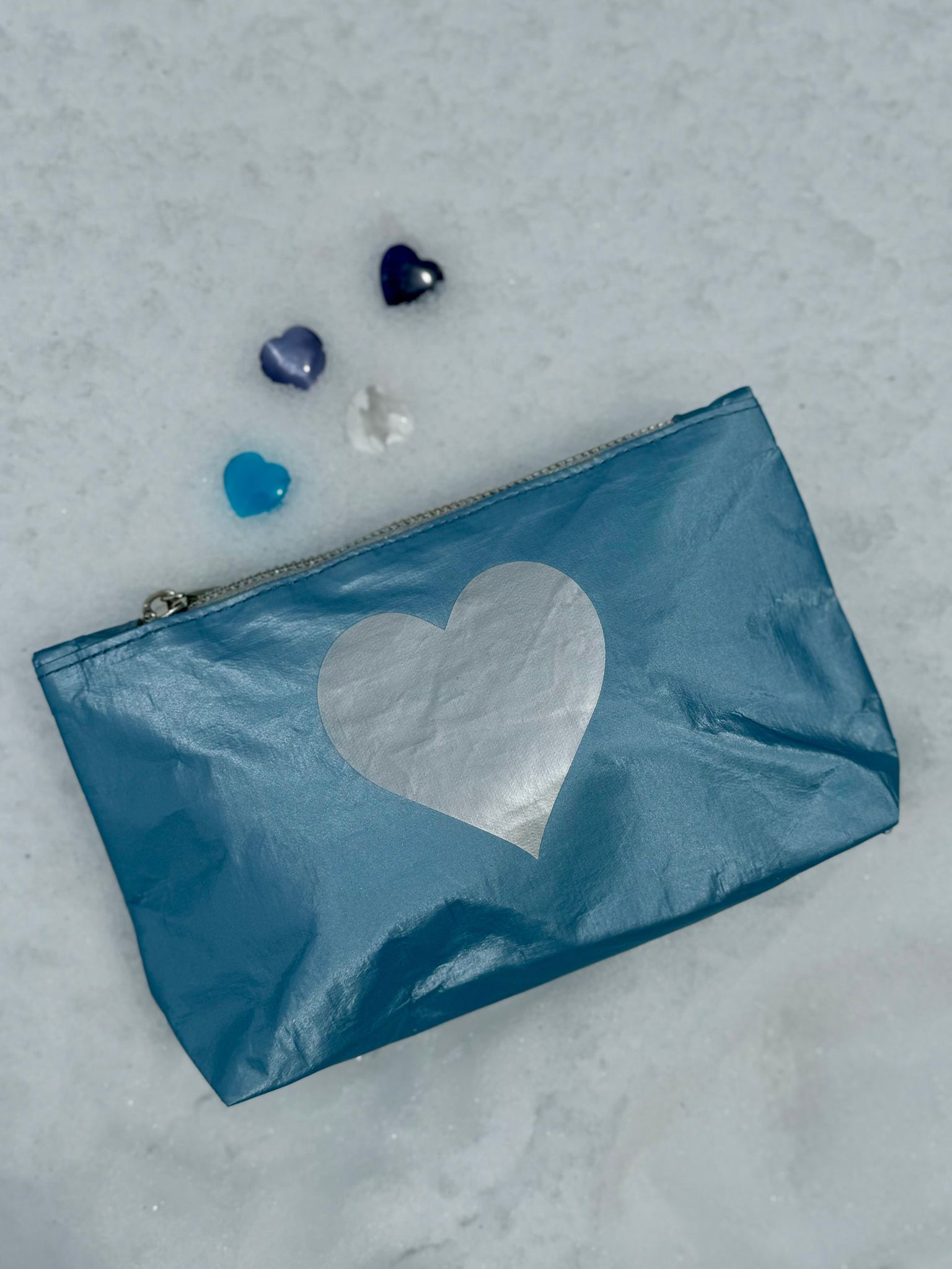 Shimmer ice blue with silver heart emblem zipper pouch laying on top of snow