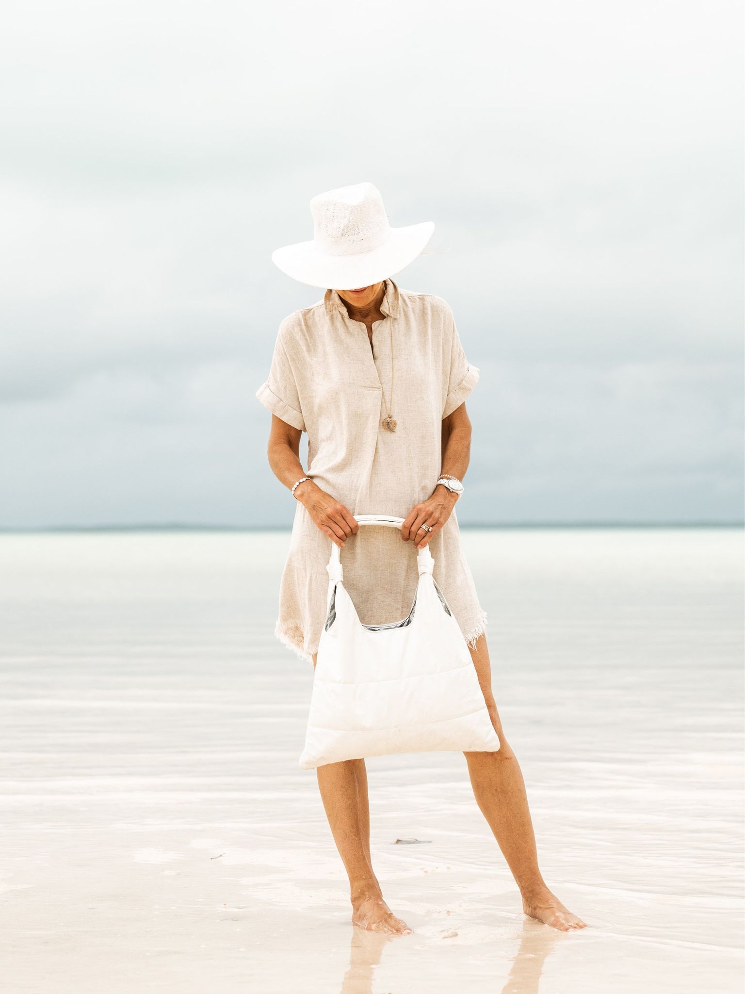 Woman in standing on ocean shoreline holding shimmer white puffer tote in front of her