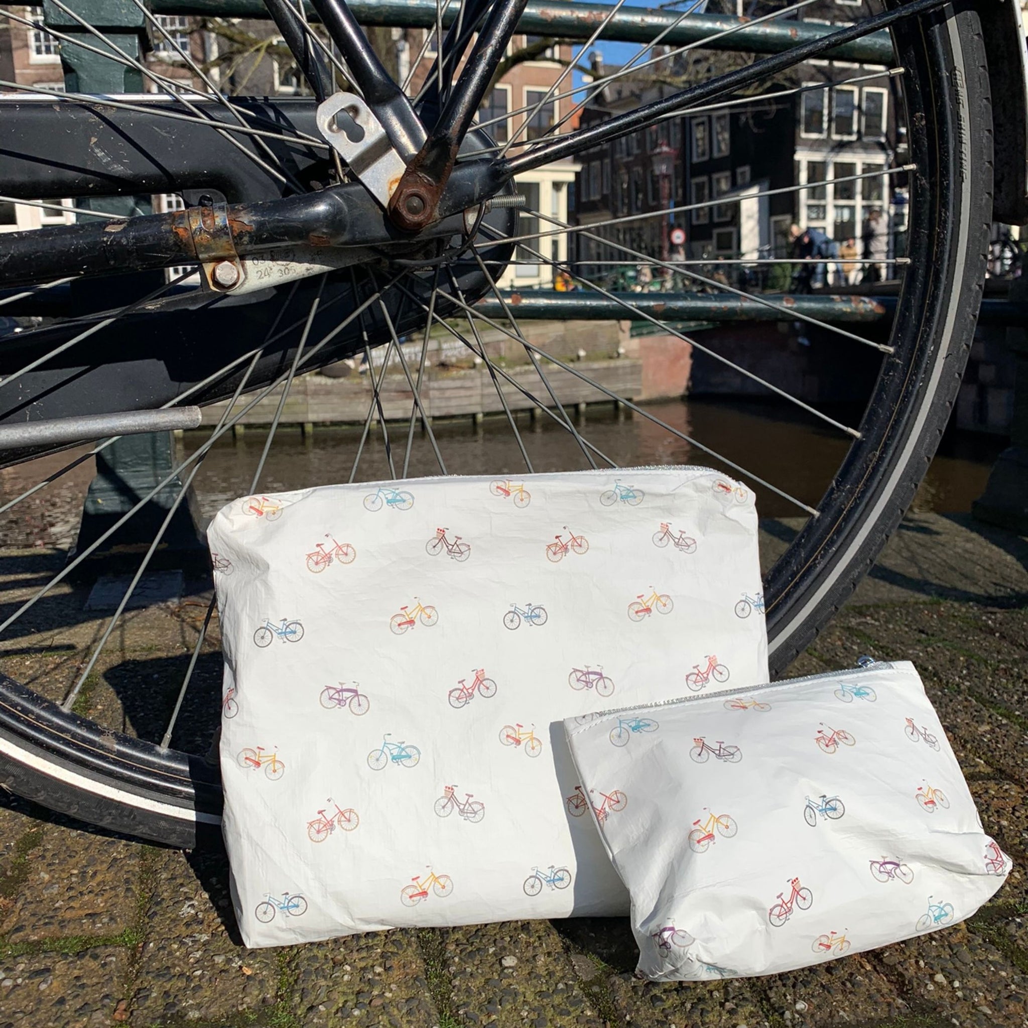 Set of two white zipper pouches with colorful bicycles pattern sitting against bicycle tire