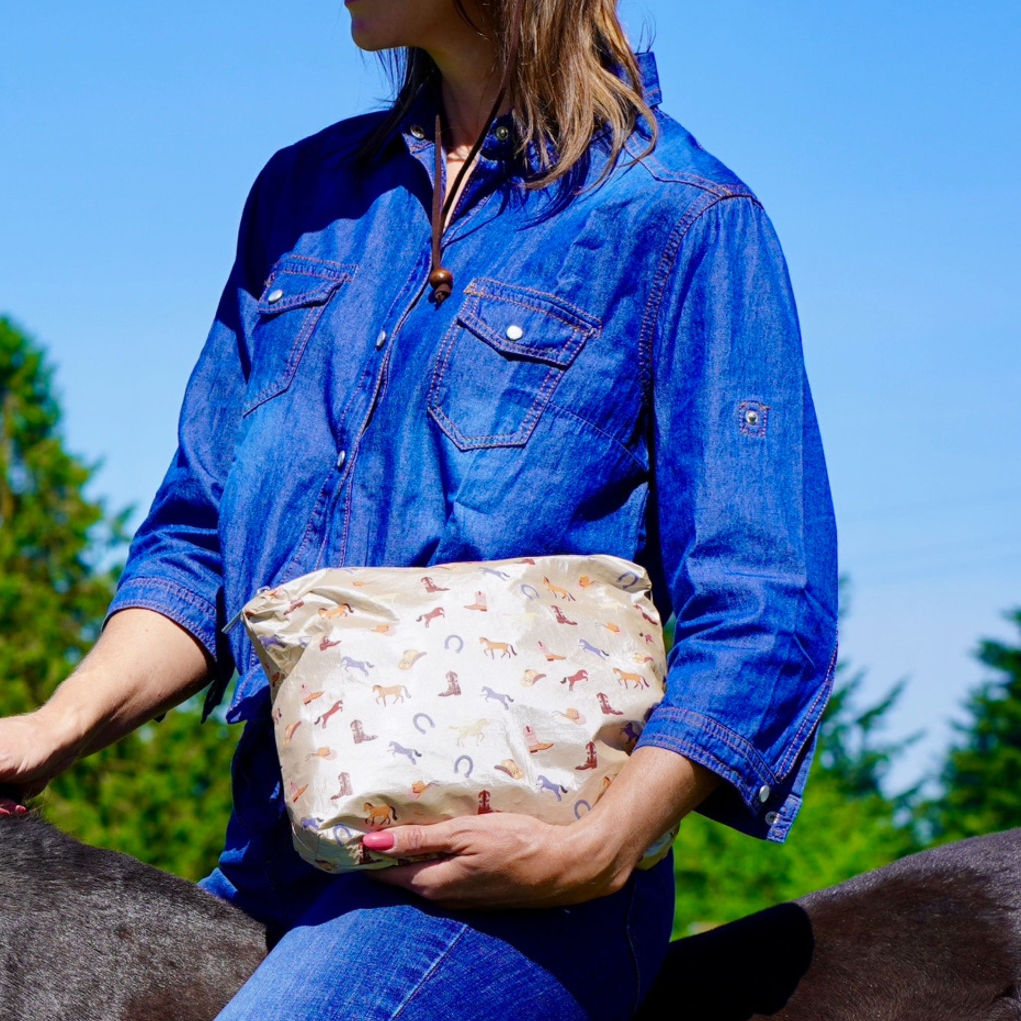 Woman holding beige clutch bag with horse, cowboy hat, cowboy boot, and horseshoe pattern